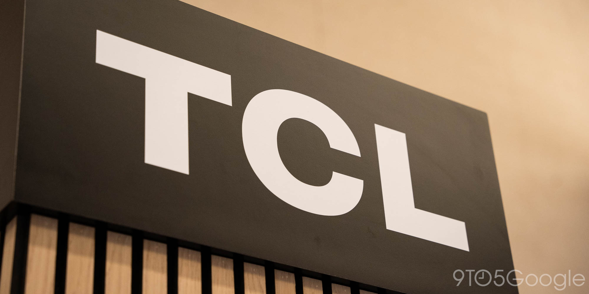لوحة إعلانية سوداء عليها شعار TCL الأبيض بخط كبير، مع إضاءة خافتة، وأسفلها جزء خشبي بخطوط عمودية، وخلفية بلون البيج الفاتح.