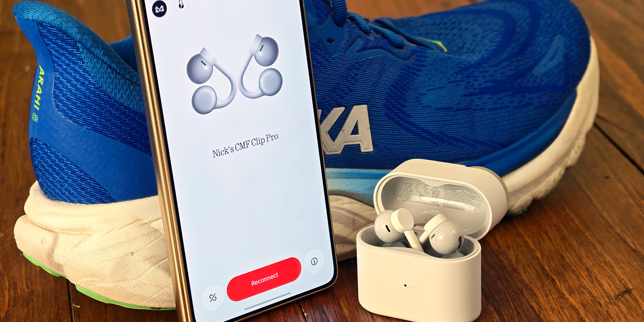 White CMF Clip Pro earbuds in their case, next to a phone with the Nothing X app and a pair of running shoes